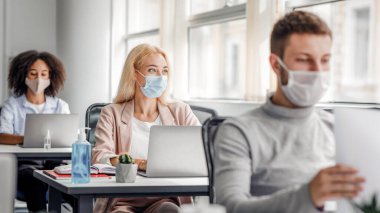 Proje üzerinde takım çalışması. Koruyucu maskeli çok ırklı çalışanlar antiseptiklerle masalarda dizüstü bilgisayarlar üzerinde çalışıyorlar, dalgın sarışın bayana odaklanıyorlar, pencereden dışarı bakıyorlar, fikir üretiyorlar, uzayı kopyalıyorlar.