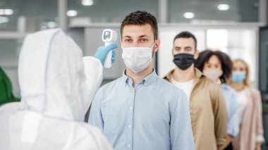 Sıcaklığı temas olmayan termometreyle belirlemek için modern teknolojiler. Tehlikeli madde giysisi giyen adam, ofise girerken koruyucu maskeler takan çok ırklı meslektaşlarının sıcaklığını ölçüyor.