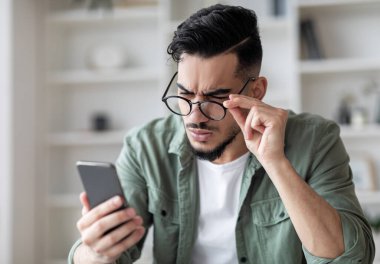 Görme sorunları kavramı. Gözlüklü genç Arap adam akıllı telefon ekranına bakıyor ve kaşlarını çatıyor, milenyum adamı mesaj okumaya çalışıyor, Astigmatizm ve kötü vizyondan muzdarip,