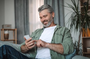 Sakallı, akıllı telefonuyla yazı yazan neşeli, yaşlı Avrupalı bir adam. Oda içinde sosyal medya sohbetinin keyfini çıkarıyor. Sosyal uzaklık ve yeni uygulama, evde bulunan emekliler için covid-19 salgını için aygıt