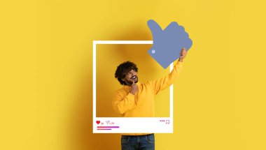 Sarı kazak giymiş neşeli bir nüfuz sahibi, beyaz çerçeveli, parlak sarı arka planda, sosyal medya platformlarında içerik oluşturma ve bağlanma için mükemmel bir başparmak verir..