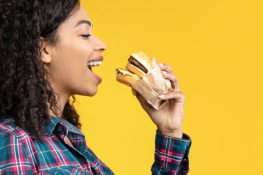 Atıştırma zamanı. Aç Siyah Kadın Hamburger Yiyen Kadın Portresi. Afro-Amerikan Kadın Fast Food 'tan ısırıldı, Orange Stüdyo Arkaplanı, Afişi, Kopyalama Alanı' nda izole edildi.
