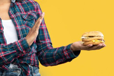 Fast Food 'u reddet. Mutsuz Afro-Amerikan kadın portresi Burger tutuyor ve Palm ile el hareketlerini gösteriyor. Siyah kadın sağlıksız sandviçi reddediyor, turuncu arka planda izole edilmiş, pankart.