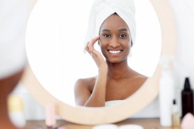 Güzellik, gençlik ve sağlık prosedürleri. Aynadaki yansıması, havluyla yüzü krem üstüne krem sürmüş, sabahları yatak odasında kozmetik ürünleriyle dolu.