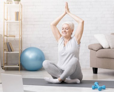 Online eğitim. Evde dizüstü bilgisayarla yoga yapan, meditasyon yapan, nefes egzersizi yapan, boş alan arayan yaşlı bir kadın.
