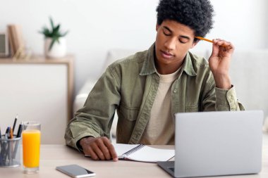 Thoughtful black teen guy passing hard exam or online test on laptop at home, panorama. Teenage male student learning remotely, doing difficult home assignment, working on educational project