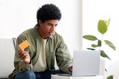 Genç siyah adam evdeki dizüstü bilgisayarında online alışveriş için kredi kartı kullanıyor. Pozitif Afro-Amerikan gençleri mal ya da market alışverişi yapıyor, internetteki hizmetlere para ödüyor