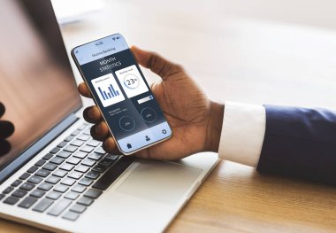 Afro-Amerikalı iş adamı cep telefonuyla bilgisayarı takas ediyor. Yaratıcı bir görüntü. Ofiste çalışırken akıllı telefondan finansal uygulama kullanan tanınamayan siyah yönetici