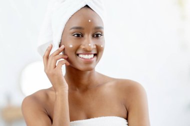 Duş sonrası rutin ve cilt bakım ürünleri. Kafasında beyaz havluyla gülümseyen Afrikalı genç bir Amerikalı kadın yüzüne krem sürüyor ve sabahları yatak odasındaki aynaya bakıyor.