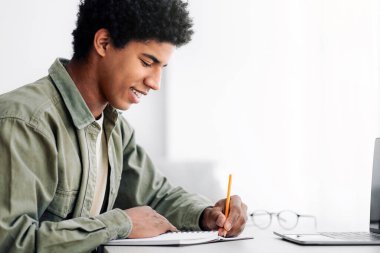 Web tabanlı eğitim konsepti. Evdeki dizüstü bilgisayarın önünde boş bir yere not defteri yazan siyah bir erkek öğrenci. Pozitif Afro-Amerikan ergen uzaktan çalışıyor, internette öğretmeniyle konuşuyor.