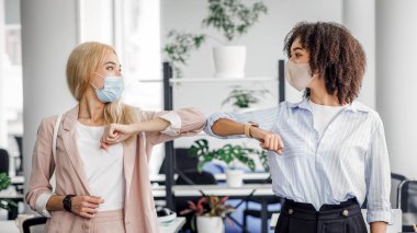 Yeni normal, koronavirüse karşı koruma ve salgın sırasında selamlama. Avrupalı ve Afro-Amerikalı koruyucu maskeli kadınlar, modern ofis içinde dirsekleriyle selam veriyorlar.