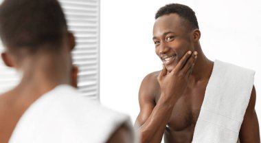 Tıraş olduktan sonra pürüzsüz yüzlere dokunan mutlu siyah adam evdeki modern banyoda aynanın yanında duruyor. Erkek yüz bakımı ve kendine bakım kavramı. Panorama