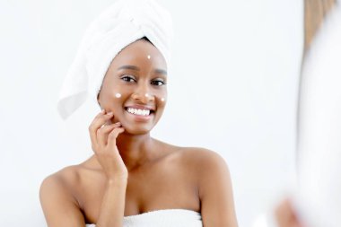 Evde spa günü ve güzellik bakımı, yalnız ve boş zaman. Binlerce yıllık mutlu Afro-Amerikan bayanı duş aldıktan sonra yüzüne krem sürüyor ve karantinadaki modern yatak odasındaki aynaya bakıyor.