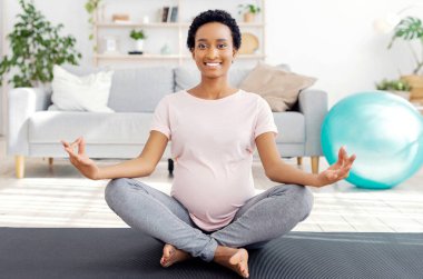 Nilüfer çiçeği pozunda yoga minderinde oturan, meditasyon yapan ya da nefes egzersizi yapan, evdeki sağlığıyla ilgilenen mutlu siyahi kadın. Güzel müstakbel anne hamilelik boyunca formunu koruyor.