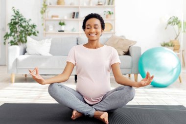Pozitif siyah hamile kadın lotus pozunda meditasyon yapıyor, gözlerini kapıyor, evde nefes egzersizi yapıyor. Mutlu müstakbel anne bebek beklerken sakin ve huzurlu.