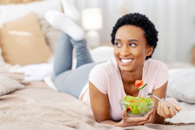Sağlıklı vejetaryen diyeti. Gülümseyen siyahi kadın evde, elinde taze sebze salatası, fotokopi odası ile yatıyordu. Afro-Amerikalı kadın vejetaryen yemeğinin tadını çıkarıyor, temiz bir yaşam için teşvik ediyor.