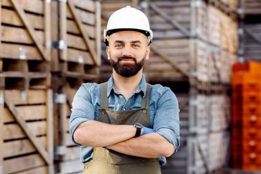 Depo, yönetim, dağıtım ve lojistikteki ürünleri inceliyorum. Üniformalı, kasketli, kolları çapraz olan mutlu, kendine güvenen genç sakallı adam, arka planda eşyalar olan tahta kutularda.