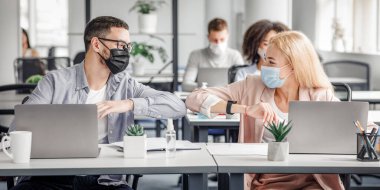 Tecritten sonra modern temassız beşlik iş başında ve yeni normaller. Mutlu milenyum erkek ve kadın koruyucu maskeli işyerinde dizüstü bilgisayarlar ve antiseptiklerle dirseklerine dokunuyor.