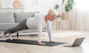Yurtiçi eğitim. Aktif kıdemli kadın evde dizüstü bilgisayarın önünde plank egzersizi yapıyor.