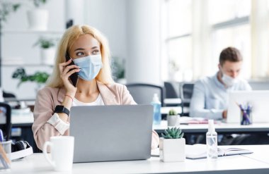 Covid-19 karantinasından sonra işine dön. Bin yıllık koruyucu maskeli bir kadın iş yerindeki bilgisayarında modern ofis içi antiseptiklerle telefonla konuşuyor, boş yer var.