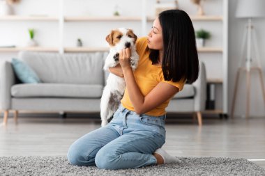 Milenyum Koreli kadın, oturma odasında halının üzerinde oturmuş, tüylü Jack Russell Terrier köpeğini tutuyor. Kadın sahibi, evinde köpeğiyle kucaklaşırken sağlıklı şeyler veriyor.