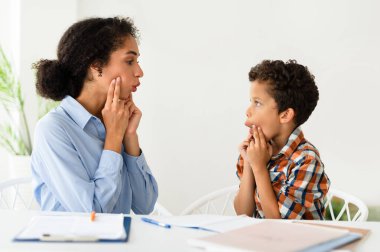 Konuşma terapisi seansı. Çocuk telaffuzu üzerinde çalışan bir kadın içeride masa başında oturuyor. Profesyonel konuşma terapisti küçük bir hastaya kekeleme konusunda yardım ediyor. Logopedik Düzeltme