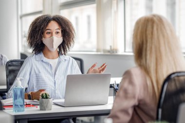 Tecritten sonra müşteri ve çalışanları bulmak. Masasında antiseptik ve dizüstü bilgisayarları olan koruyucu maskeli Afrikalı Amerikalı bir kadın modern ofis içlerindeki bir bayanla konuşuyor. Salgın sırasında, boş alan.