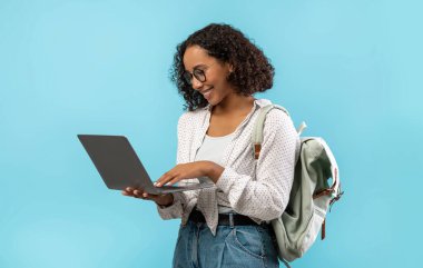 Sırt çantalı mutlu, genç Afrikalı Amerikalı kız öğrenci mavi stüdyo arka planında dizüstü bilgisayar kullanıyor. Uzaktan eğitim, çevrimiçi eğitim, öğrenim kavramı