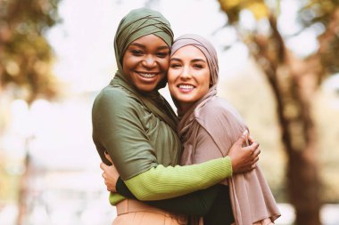 İki Müslüman kadın tesettüre sarılmış, kameraya gülümsüyor dışarıda parkta dikiliyor. Kadın Arkadaşlığı ve Birliktelik Konsepti. Siyah ve Beyaz Müslüman Kadınlar kucaklaşıyor. Panorama, Boşluğu Kopyala