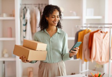 Bir elinde paketler, diğer elinde akıllı bir telefon olan neşeli genç girişimci, arka planda elbise askılarıyla şık bir ofiste duruyor.