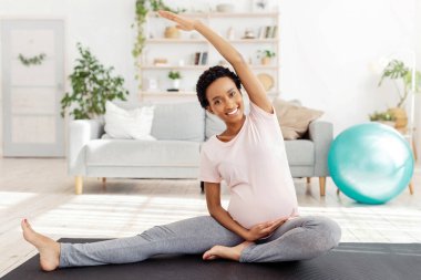 Aktif hamilelik kavramı. Gülümseyen siyah anne bacaklarını esnetiyor, sabah yoga egzersizini evde spor salonunda yapıyor. Ev içi spor yapan, ev içi spor yapan, hamile bir kadın.