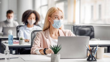 Covid karantinadan ofise döndükten sonra modern ofis çalışanı. Bin yıllık koruyucu maskeli, akıllı saatli, dizüstü bilgisayarlı bir kadın pencereye bakar ve fikir üretir.