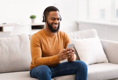 Kulaklık takan, akıllı telefon kullanan, internetten müzik dinleyen ya da evdeki koltukta oturan siyah adam. Telefon Kullanıcısı Kablosuz Kulaklıklardaki Sesin Tadını Çıkartıyor