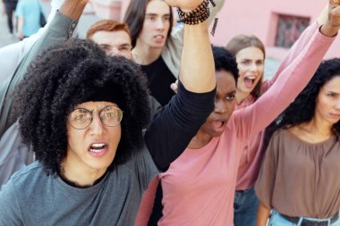 Uluslararası göstericilerden oluşan bir grubun sokakta grev yaptığını, slogan attığını ve yumruğunu kaldırıp yaklaştığını görüyoruz. Hükümet politikalarını protesto eden çok ırklı kadın ve erkekler