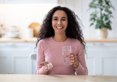 Evdeki mutfaktaki masada oturan ve elinde bir bardak su ve güzellik ilacı tutan mutlu esmer kadın. Milenyum Gülümseyen Kadın Sağlıklı Dokunulmazlık veya Güzel Cilt İçin Vitamin Hapları Kullanıyor