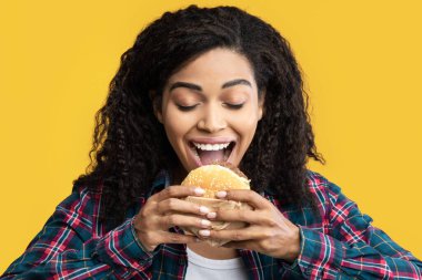 Fast Food Bağımlısı. Aç Kara Kadın Modeli Isıran Burger 'in Yakın Çekim Portresi. Kıvırcık saçlı ve lezzetli atıştırmalıklar yiyen, turuncu arka planda izole edilmiş Afro-Amerikalı bir kadın.