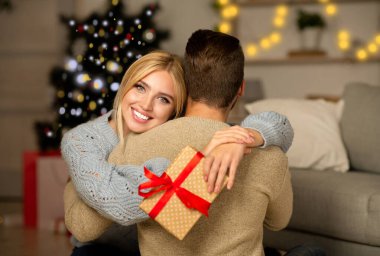 Mutlu bir eş, elinde Noel hediyesiyle kocasına sarılıyor, Noel 'i evde birlikte kutluyor, boş bir alanda. Genç bir kadın erkek arkadaşından Noel hediyesi aldığı için mutlu, onu kucaklıyor ve gülümsüyor.