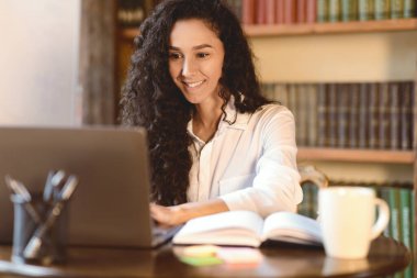 Çalışma alanı kavramı. Gülümseyen kıvırcık kadının portresi bilgisayarda çalışır ve kullanır, dizüstü bilgisayarda klavyede yazar. Masasında oturan genç bayan açık bir defter ve bir fincan kahve, bulanık bir arka plan.
