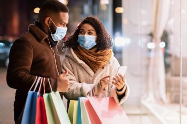 Perakende, Teknoloji ve Salgın Konsepti. Tıbbi maske takan, akıllı telefon kullanan, ekranı işaret eden ve elinde alışveriş torbaları olan kocasına uygulama gösteren Afrikalı Amerikalı bir çiftin portresi.