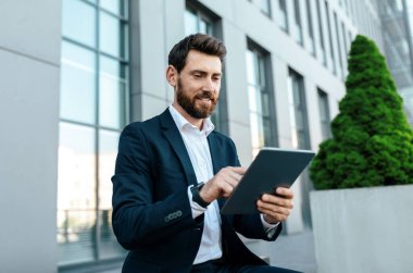 Smiling young handsome european businessman with beard in suit typing on tablet near modern office building, outdoor. Chat in new app, manager work with technology and new normal, business in city