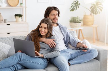 Aile hafta sonu konsepti. Kucaklaşan genç evli çift, televizyon izleyen adam ve eş dizüstü bilgisayar kullanıyor, ev içi, boş alan. Erkek ve kadın birbirlerine aşıklar oturma odasında, birlikte zaman geçiriyorlar.