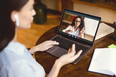 Çevrimiçi Kurs ve Uzaktan Eğitim Konsepti. Kulaklık takan, İngilizce öğretmeniyle çevrimiçi video görüşmesi yapan, konuşan, el kol hareketi yapan, bilgisayar kullanan kulaklıklı genç bir kadının omuzlarının arkasında.