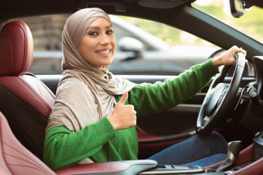 Ehliyet kursu. Orta Doğulu mutlu bir kadının portresi başparmaklarını göstererek lüks bir arabanın içinde oturuyor. Gülümseyen Müslüman Kadın Müşteri Onaylama Hareketi yapıyor, Ehliyetini aldı