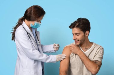 Aşı kavramı. Erkek hastalara aşı yapan şırıngalı kadın doktor, Kovan 19 'a karşı aşılanan gülümseyen genç adam, Mavi Arkaplan' da otururken,