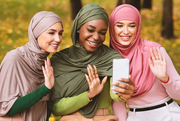 Akıllı telefonlu üç modern Müslüman kadın parktaki bankta oturmuş el sallayarak selam veriyor. İslami Öğrenciler Telefondaki Görüntü Konferansı, Uzaktan İletişim Konsepti