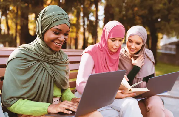 Üç modern Arap kadın öğrenci dışarıda park yerinde dizüstü bilgisayar ve kitaplarla birlikte oturarak öğreniyor. Çevrimiçi Üniversite Eğitimi Müslüman Kadınlar İçin Çevrimiçi Çalışma. Seçici Odaklanma