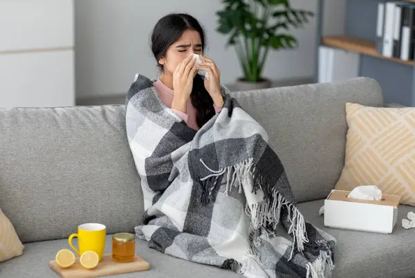 Mutsuz genç Hintli bayan peçeteye hapşırıyor, burnunu siliyor, ateşi var, kanepede yatıyor çay, bal ve limonla birlikte. Öksürük, akıntı, soğuk ve solunum yetmezliği için ev tedavisi.