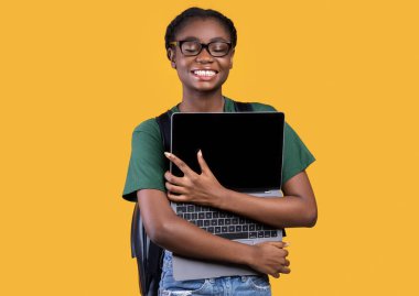 Stüdyoda sarı arka planda dizüstü bilgisayarı tutan ve kucaklayan mutlu siyah kız öğrenci. Çevrimiçi Çalışma, Eğitim Malzemeleri ve Teknoloji, Modern Yaşam Tarzı Konsepti