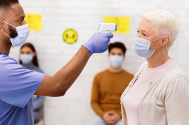 Doktor randevusundan önce yaşlı bayan hastaya ateş ölçümü yapan hemşire temassız kızılötesi termometreyi yakın mesafede tutuyor. Coronavirüs Salgını