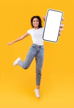 Güzel genç bir bayan yeni mobil uygulama öneriyor, dikey kolaj. Akıllı telefonu tutan heyecanlı bayan, beyaz boş ekranı gösteriyor, turuncu sarı stüdyo arka planında zıplıyor, tam boy